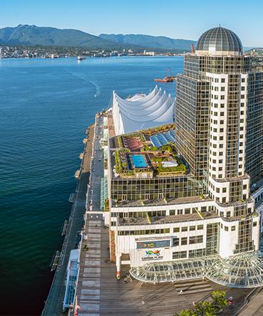 Vue aérienne de la façade du Pan Pacific Hotel Vancouver, avec le port de Vancouver et les montagnes en arrière-plan. 