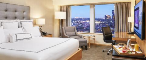 A deluxe room at the Pan Pacific Hotel Vancouver includes a king-size bed with headboard, a nightstand with lamp and telephone on it, a chair with ottoman, side table with an overhead lamp, and a desk with a chair. A skyline view of Vancouver is visible through the window